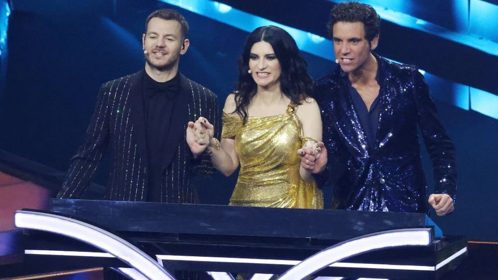 The three hosts of Turin 2022 anxiously hold hands while waiting for vote results