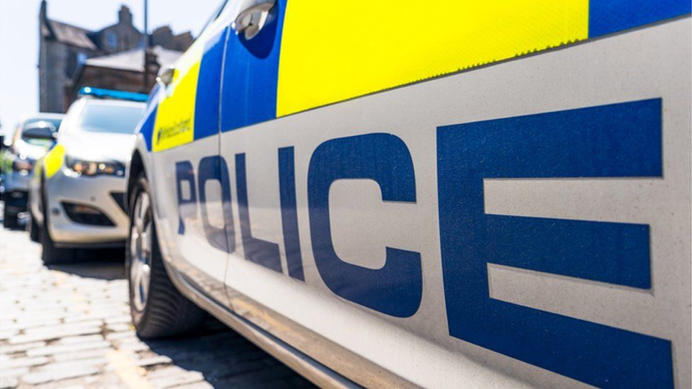 The word 'Police' below the fluorescent decals on a police car on a UK street, with other police vehicles in the distance.