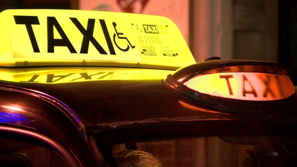 An illuminated taxi sign on top of a black cab