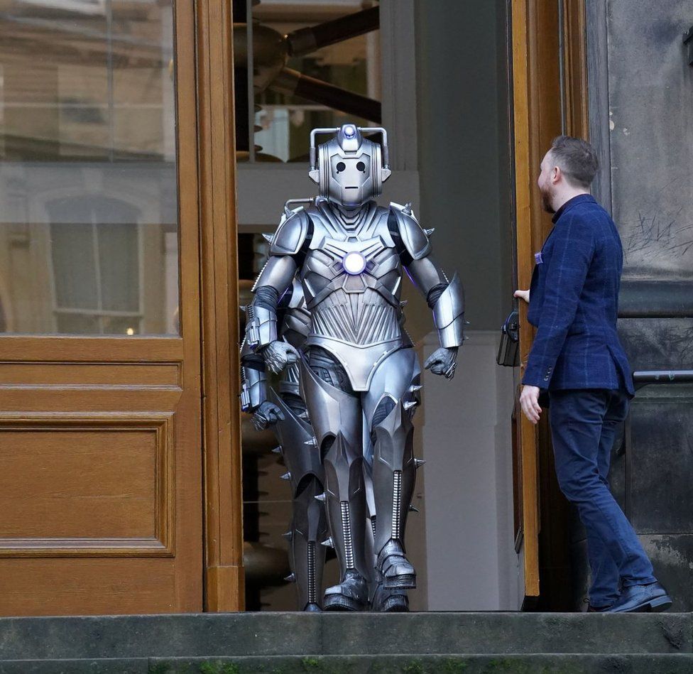 A curator opens a museum door for a cyberman