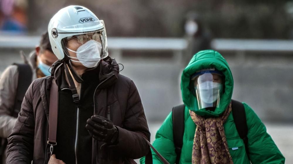 A man wearing a helmet and n95 face mask walks through the streets and a woman wearing a mask and faceshield behind him