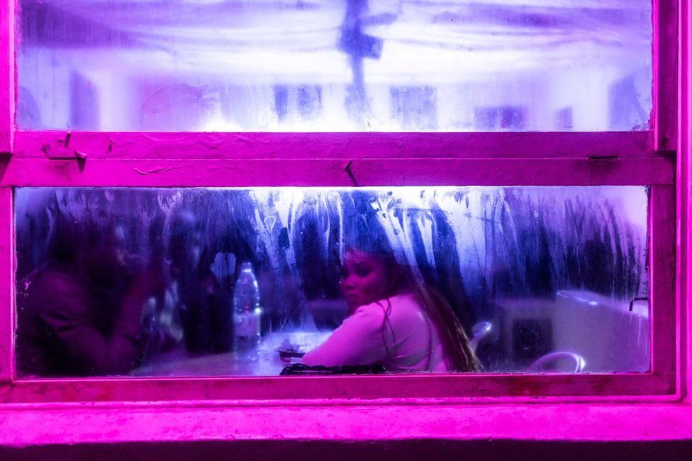 A woman looks out of a dark bar that is surrounded in neon pink lighting.