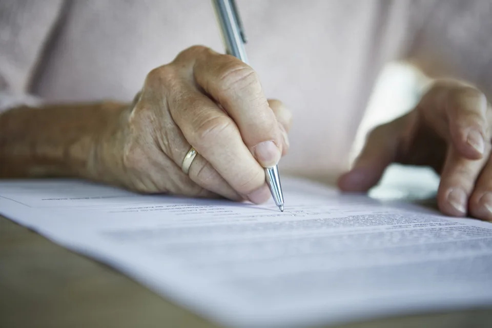 A close-up of an elderly person’s hand signing a document with a pen, wearing a simple ring. The article focuses on subjects related to work and money