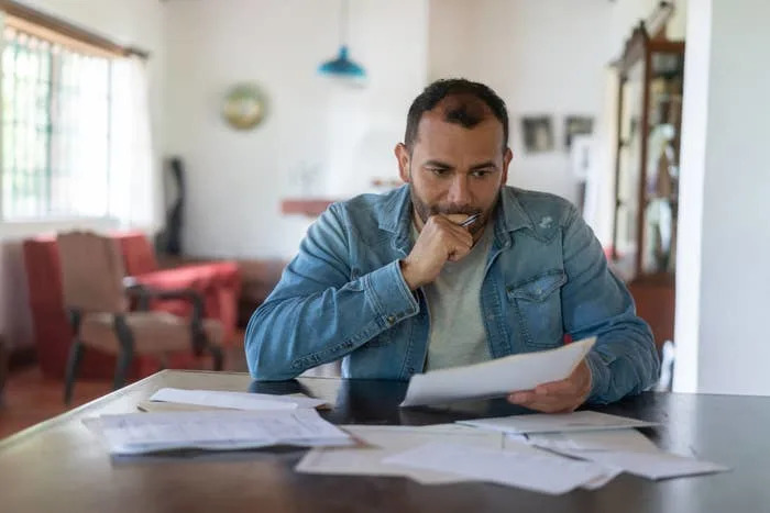 A man in a denim jacket sits at a table with various papers and a pen in his hand, looking concerned. The scene suggests he is handling financial documents