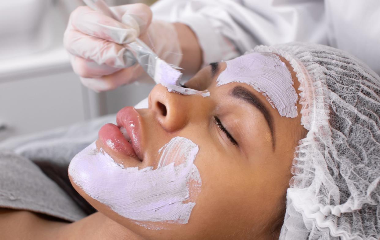 Cosmetology. Cosmetologist applies a cleansing mask on black woman face with a brush. Cosmetic procedure for facial rejuvenation.